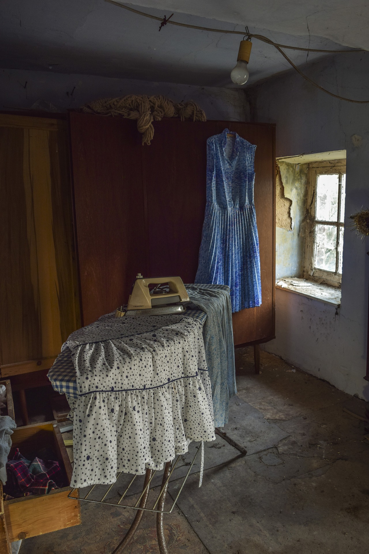 uma tábua de passar a ferro com roupa em cima num quarto que aparece abandonado